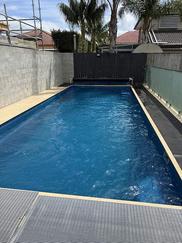 Long lap pool with a pool cover roller and glass balustrade beside a dark timber fence